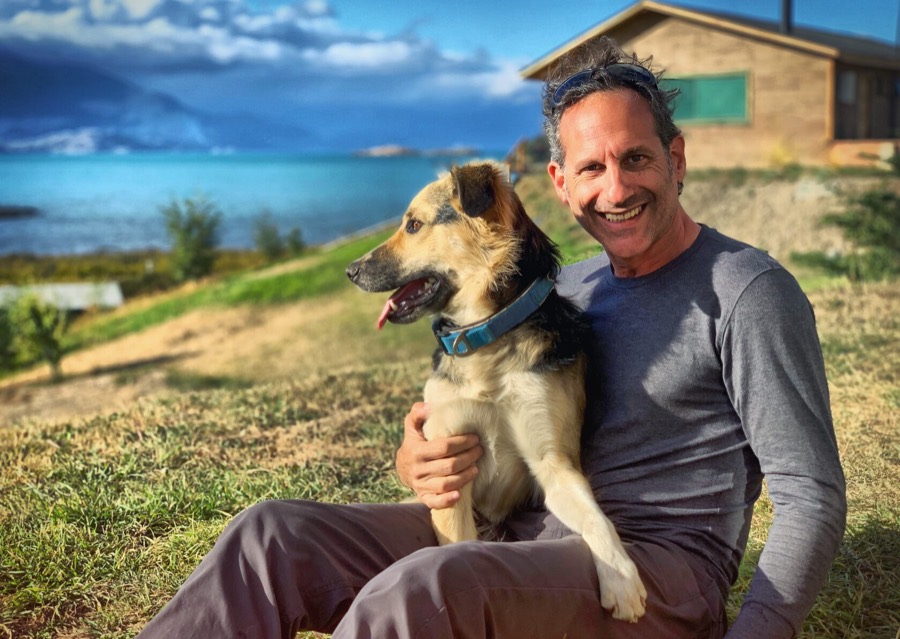 Steven on the grass hugging a dog, with a lake and mountains behind