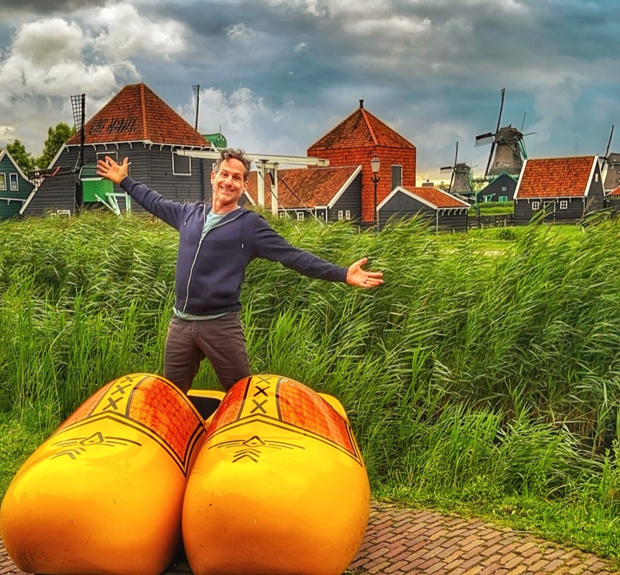 Steven with arms spread behind giant wooden clogs, with windmills behind