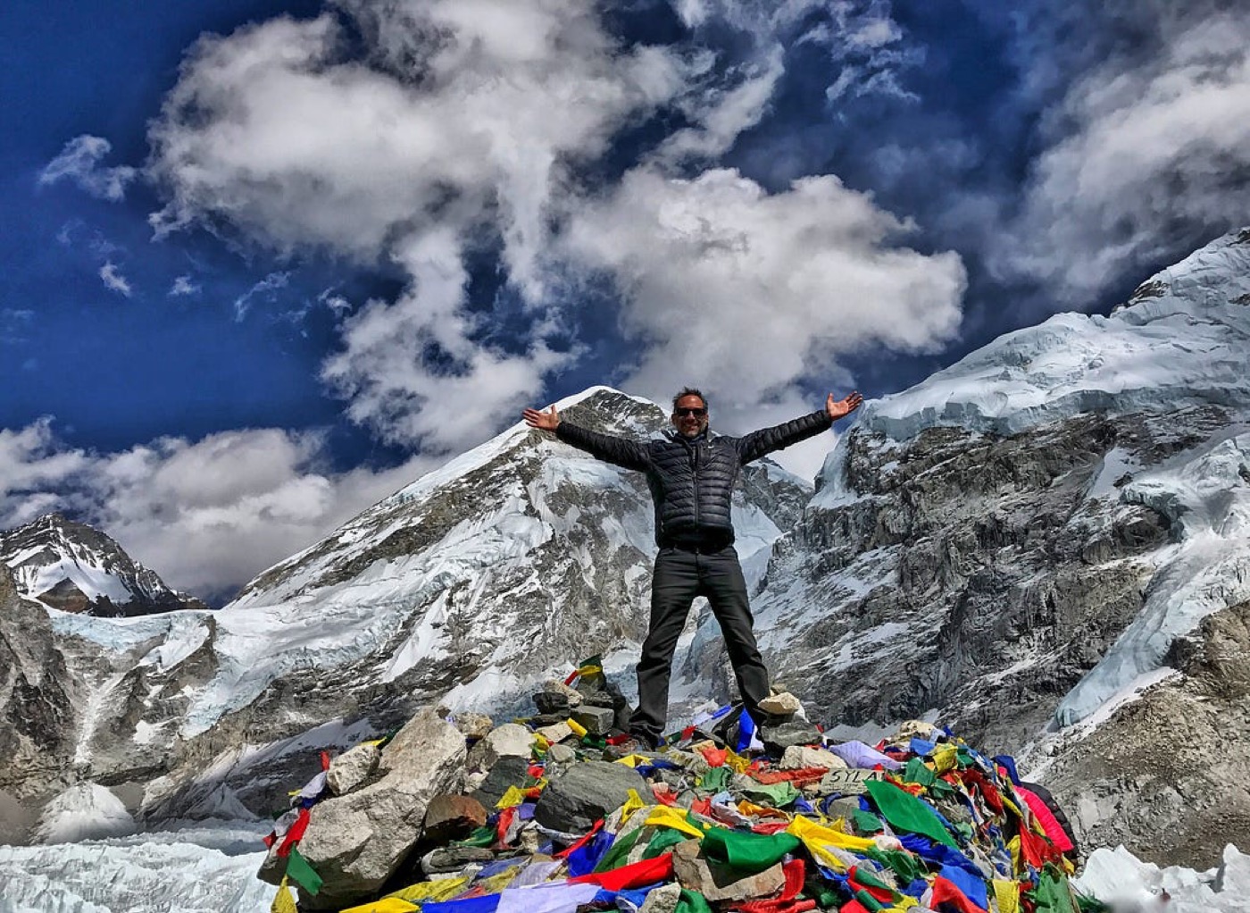 Steven with arms raised on a pile of rocks and prayer flags, glaciers and peaks behind him