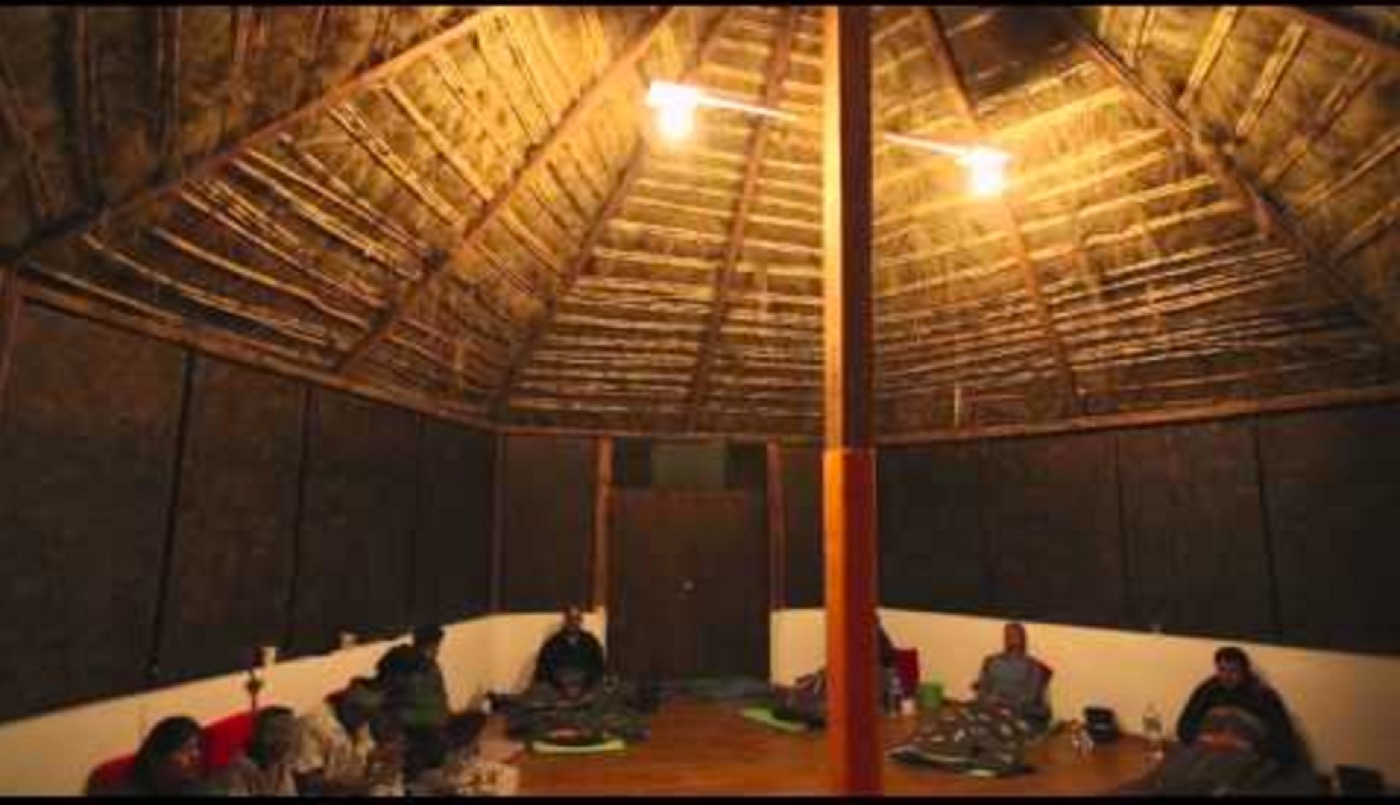 The inside of a round thatched-roof ceremonial hut at night, people sitting around its edge