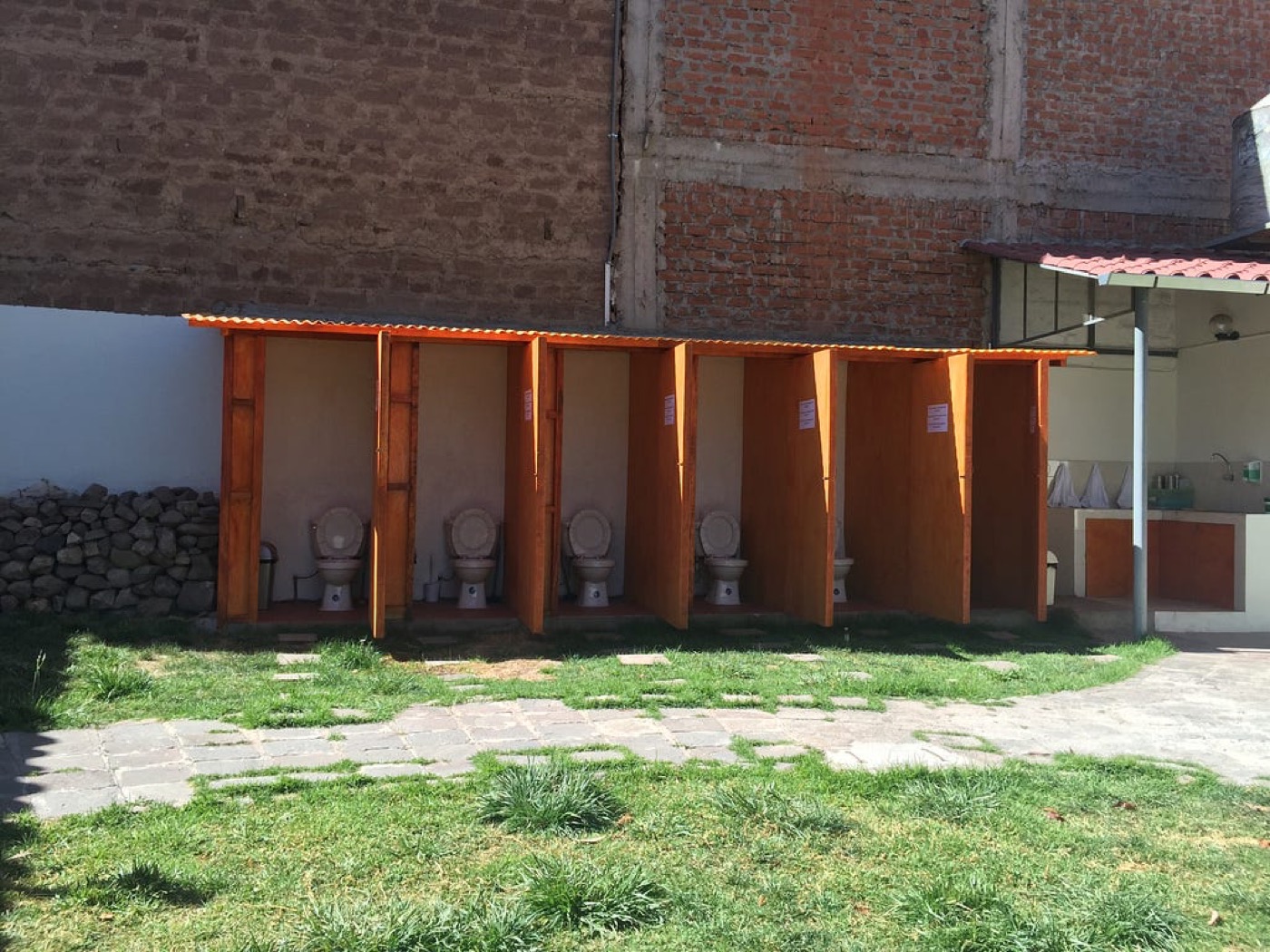 A row of outdoor toilet stalls with orange wooden walls in a grassy courtyard