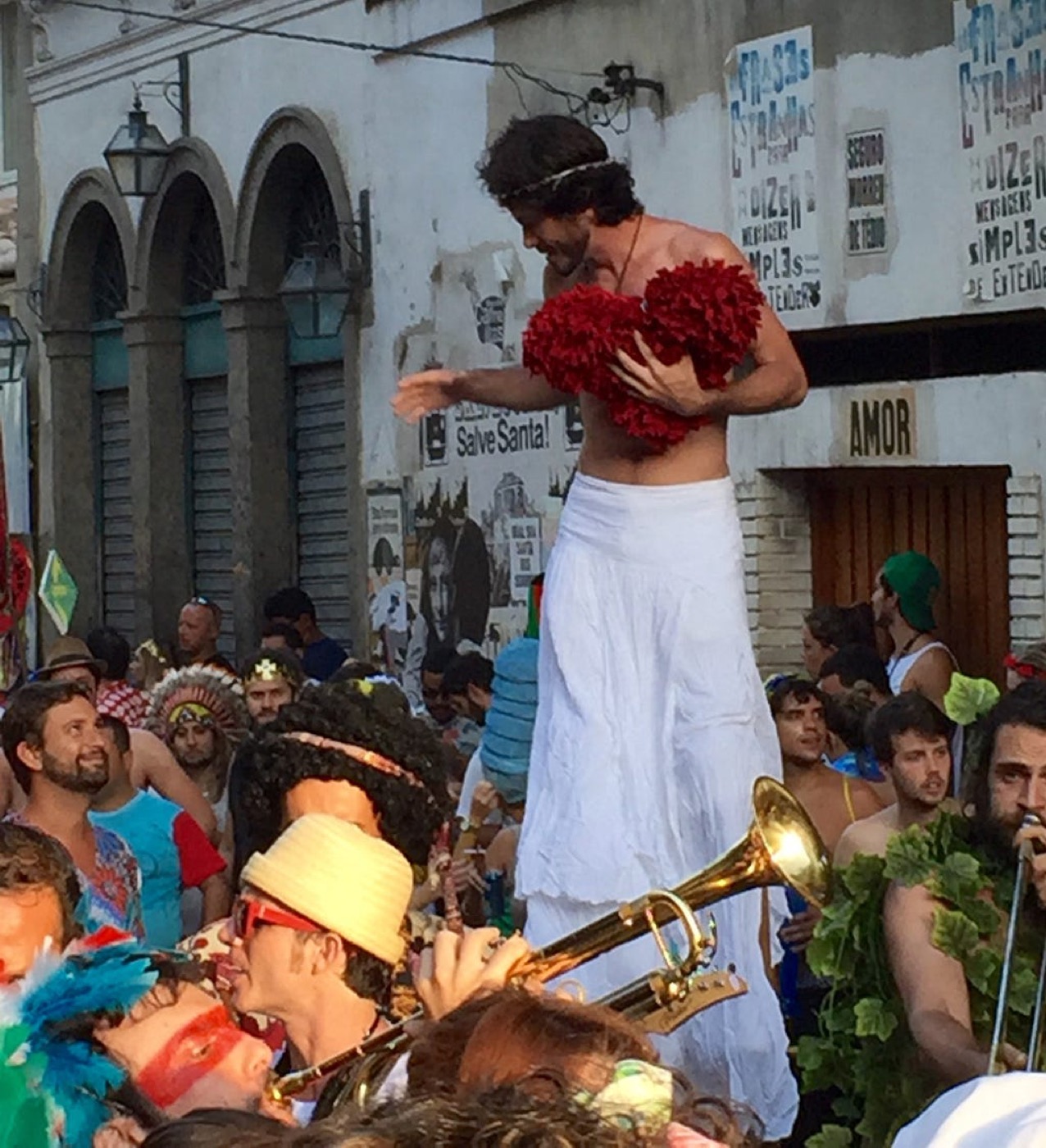 A street performer dressed as Jesus with a red heart-shaped pillow, above a Carnaval crowd and brass band